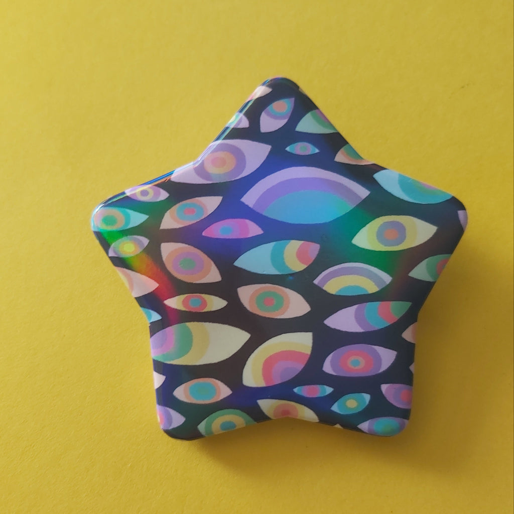 A star-shaped pin or badge featuring a colorful psychedelic pattern of stylized eyes on a dark background, set against a solid yellow backdrop.