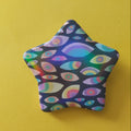 A star-shaped pin or badge featuring a colorful psychedelic pattern of stylized eyes on a dark background, set against a solid yellow backdrop.