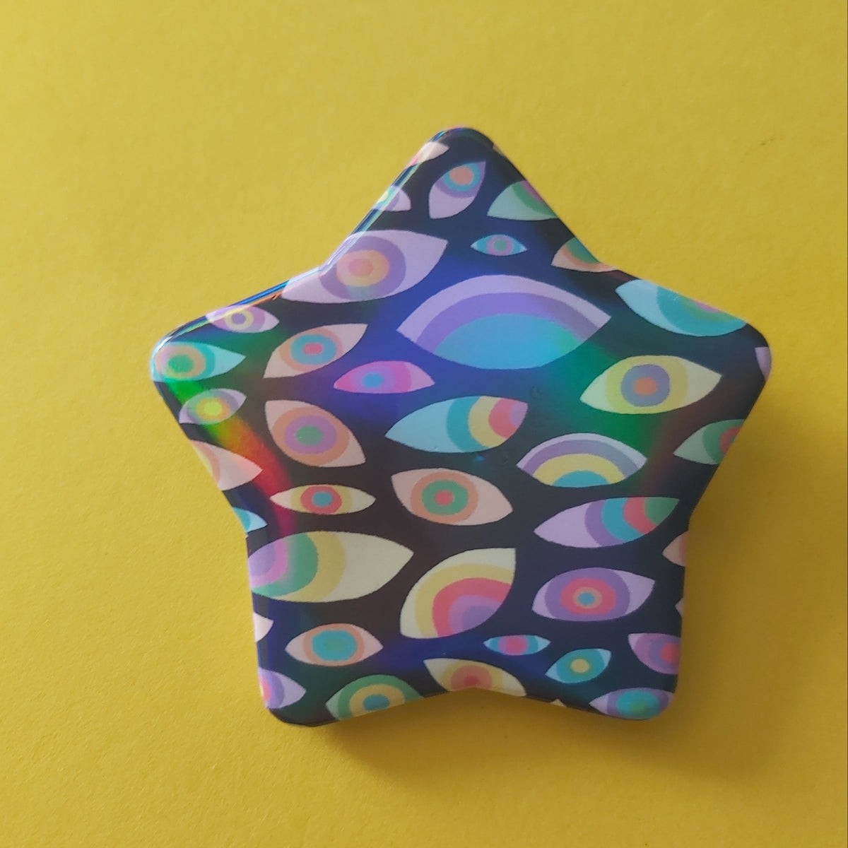 A star-shaped pin or badge featuring a colorful psychedelic pattern of stylized eyes on a dark background, set against a solid yellow backdrop.