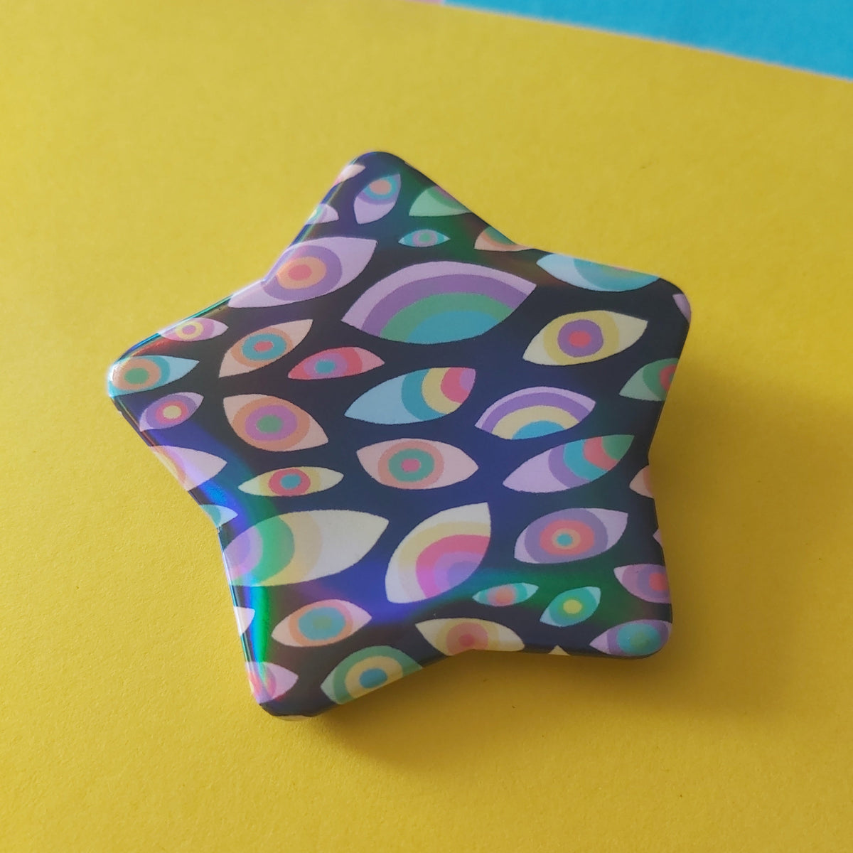 A star-shaped pin with a colourful psychedelic eye pattern on a dark background, set against a bright yellow paper backdrop with pastel colored corners.