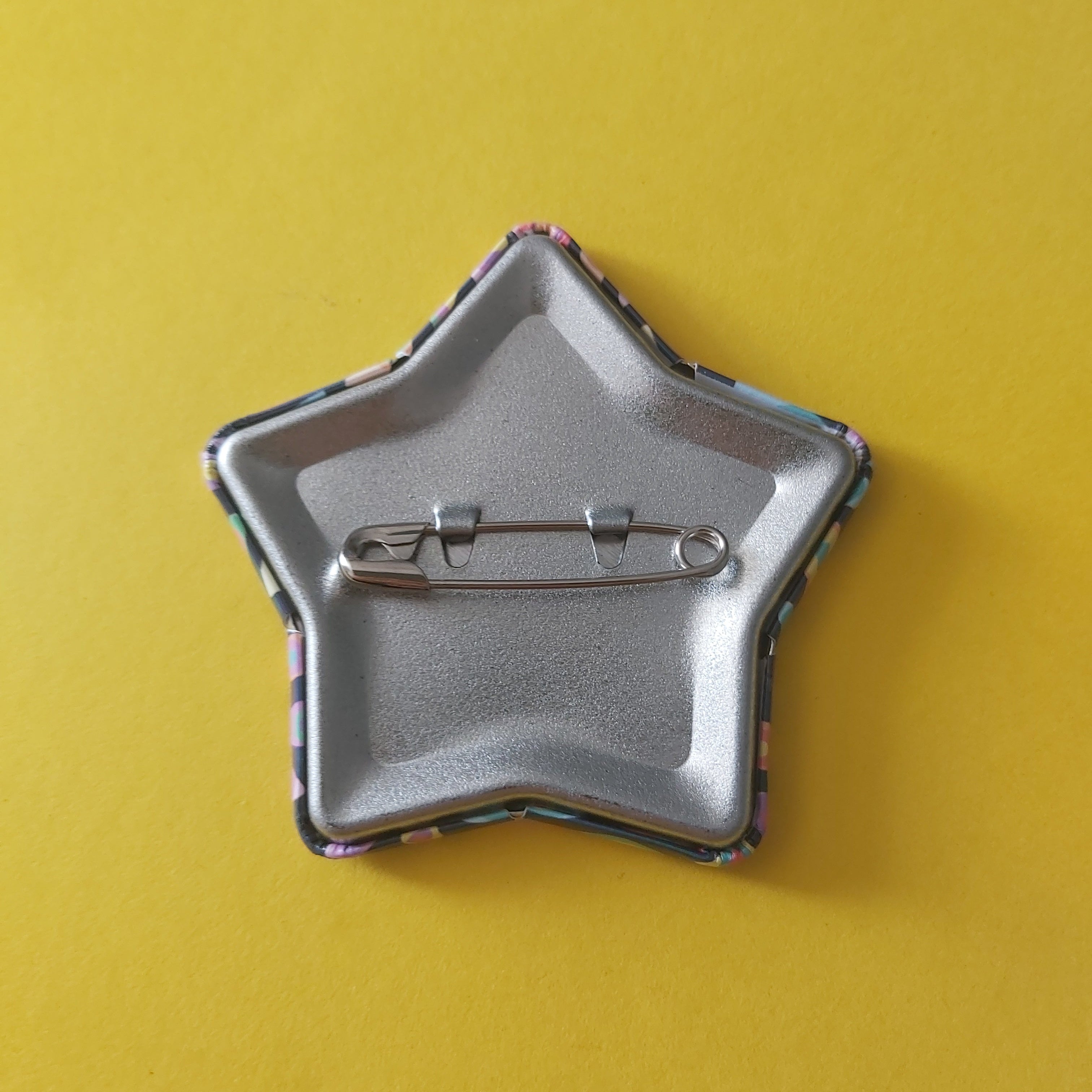 The back of a star-shaped pinback button with a silver metal base and a colourful patterned border, shown on a solid yellow background.