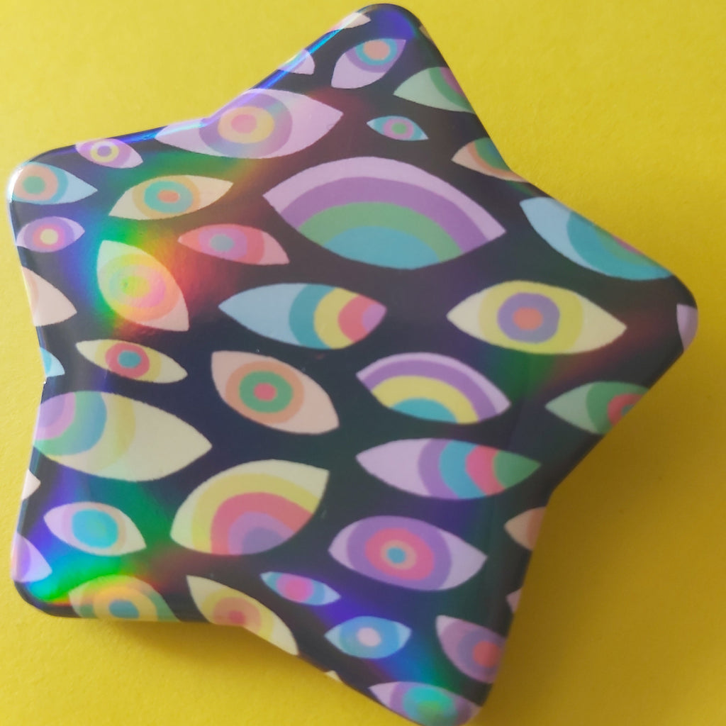 A star-shaped pin badge with a colourful psychedelic pattern of rainbow eyes and arches on a dark background, set against a solid yellow backdrop.