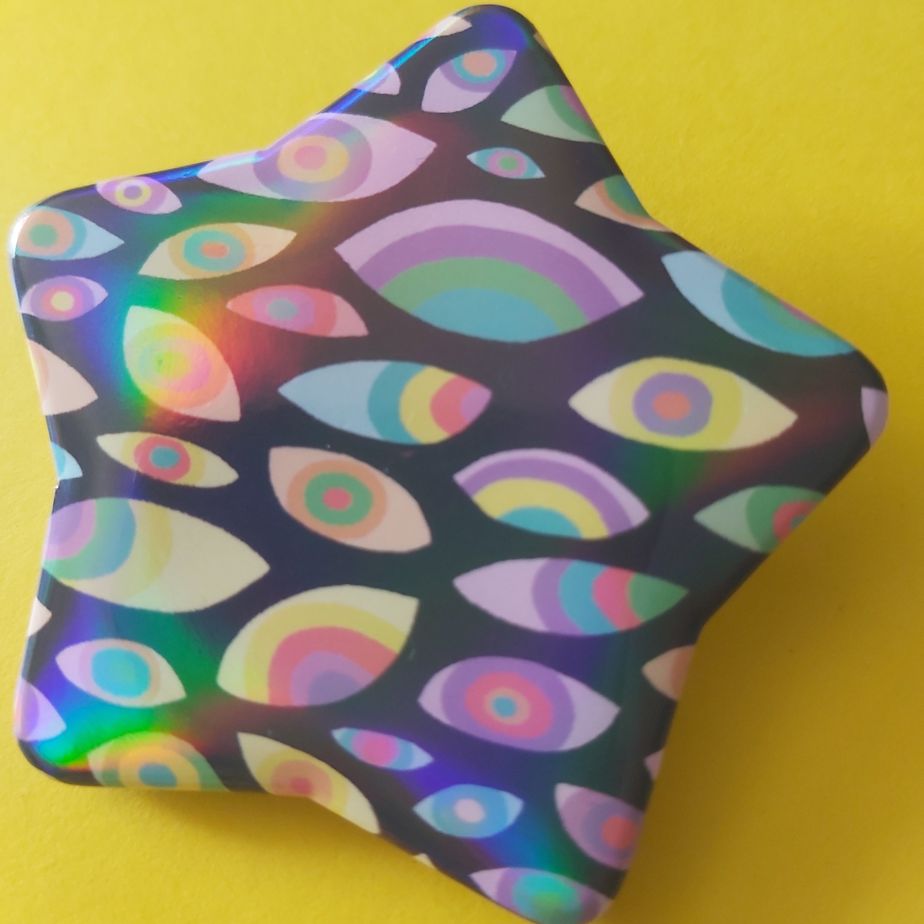 A star-shaped pin badge with a colourful psychedelic pattern of rainbow eyes and arches on a dark background, set against a solid yellow backdrop.