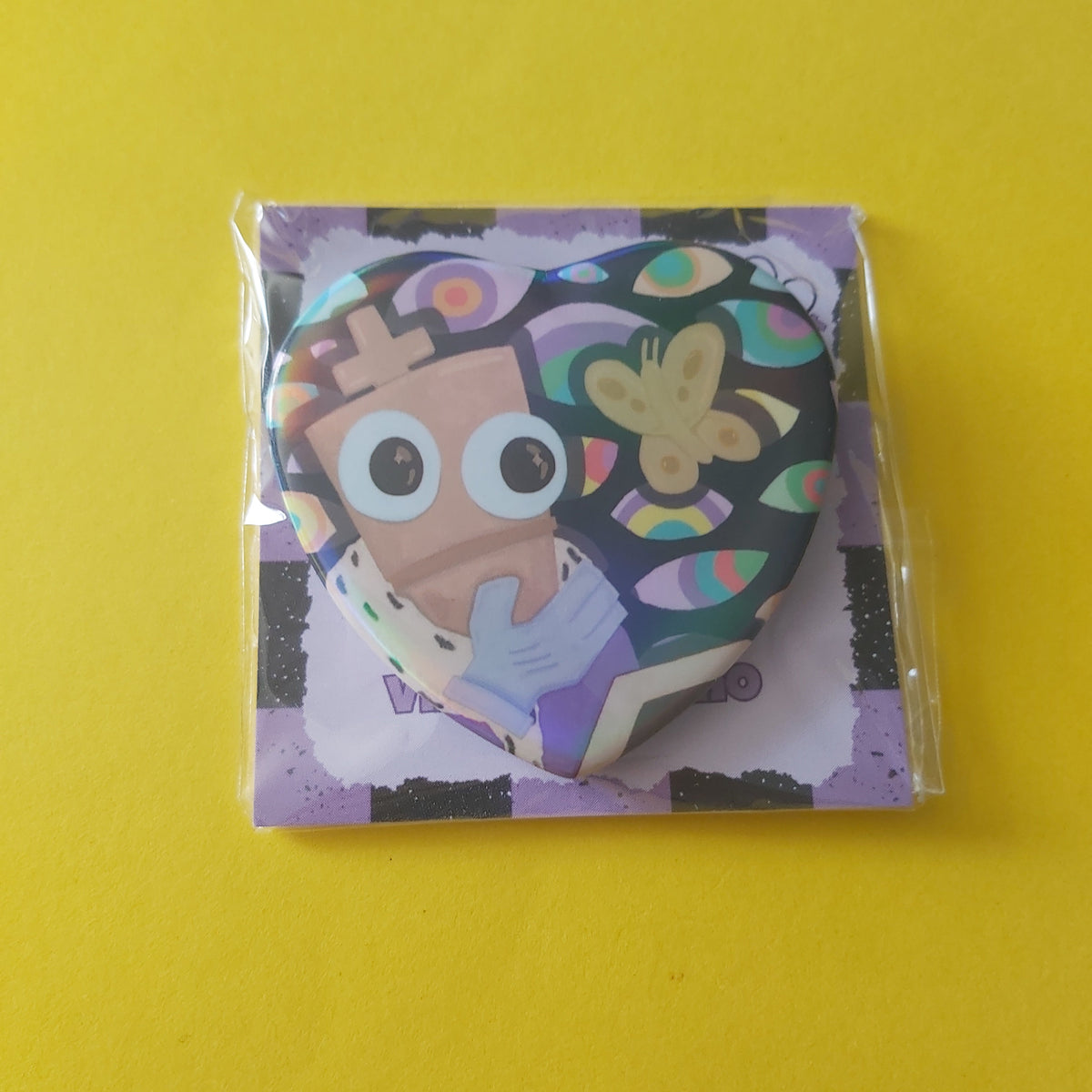 A heart-shaped pin featuring a colorful cartoon character with large eyes, packaged in a clear plastic sleeve on a purple backing card, set against a solid yellow background.