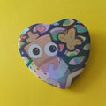 A heart-shaped pinback button featuring a colorful cartoon illustration of a character with large eyes and a butterfly, set against a solid yellow background.