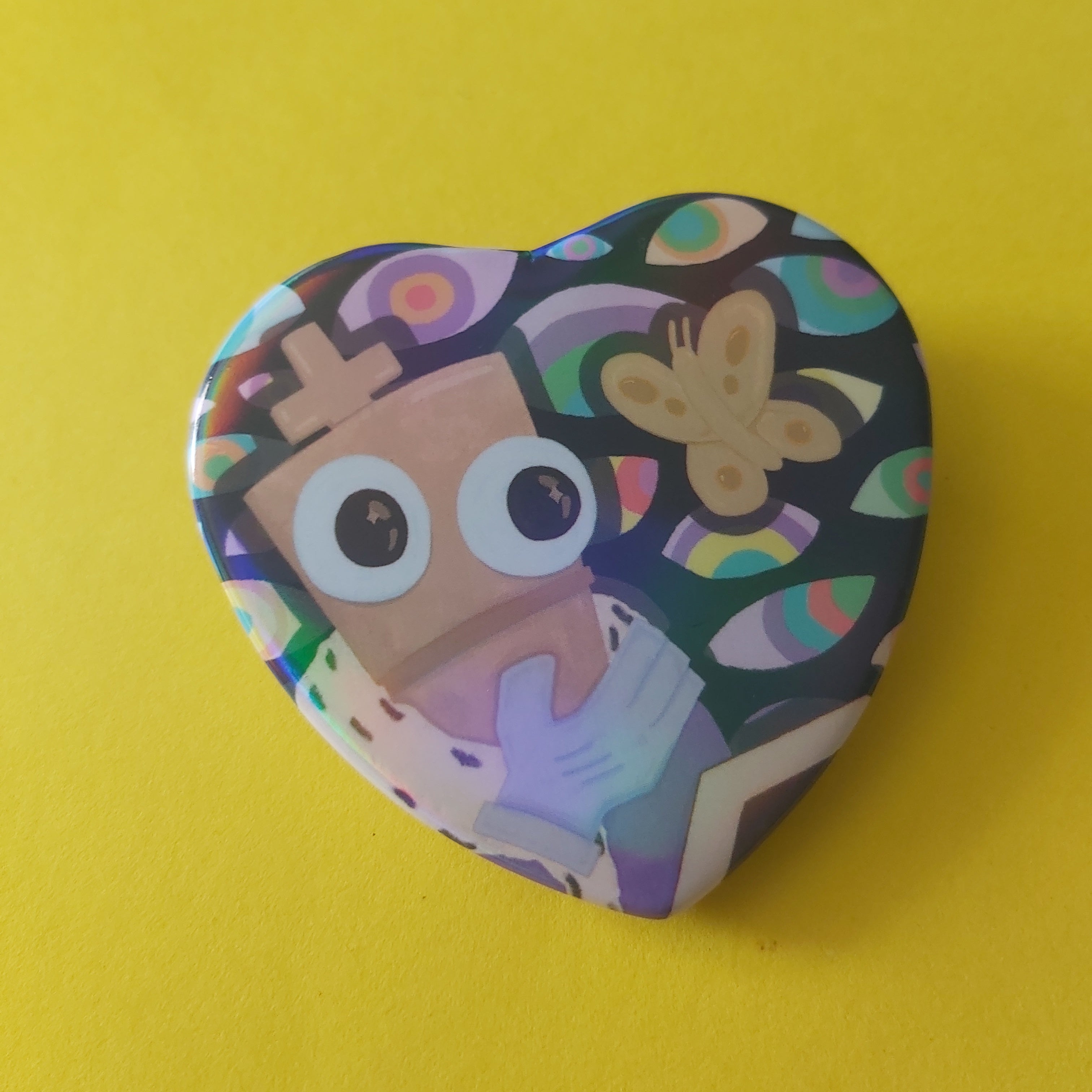 A heart-shaped pinback button featuring a colorful cartoon illustration of a character with large eyes and a butterfly, set against a solid yellow background.