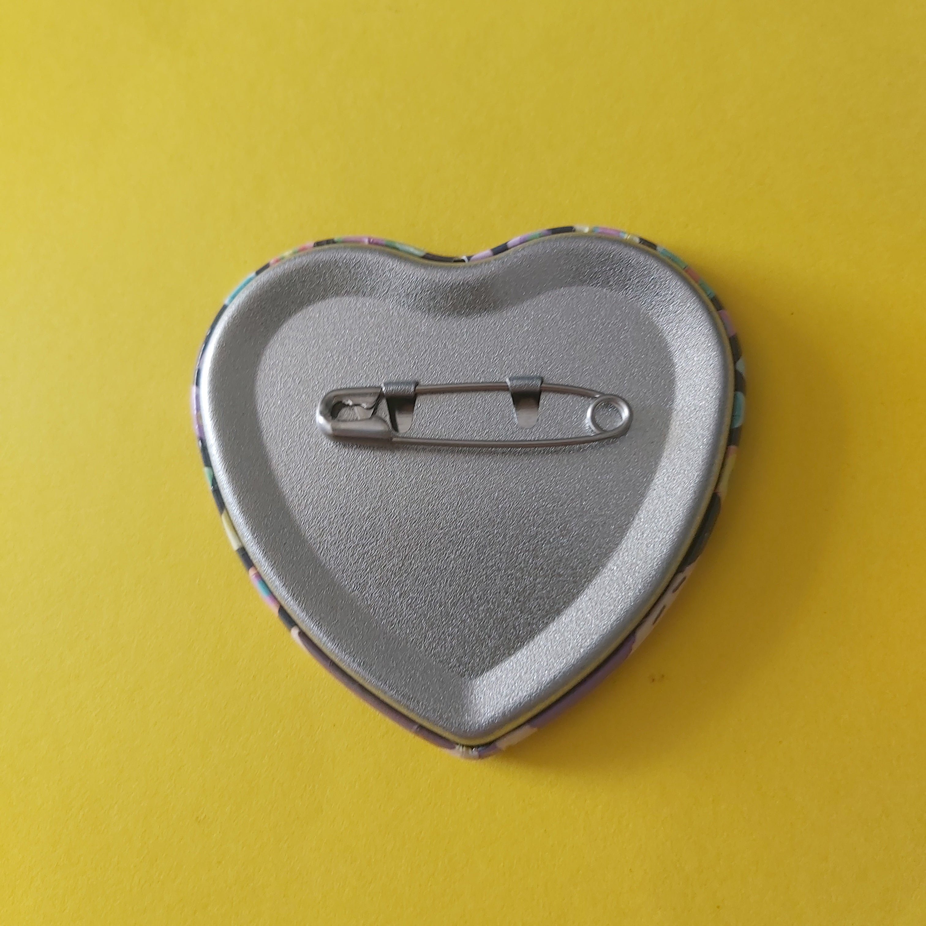 A heart-shaped pinback button showing its silver metal safety pin back, set against a solid yellow background.