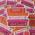 A pile of die-cut stickers featuring stacked books in orange, pink, and red with the words 'YURI', 'Sapphic', and 'Girl Love' written on them.