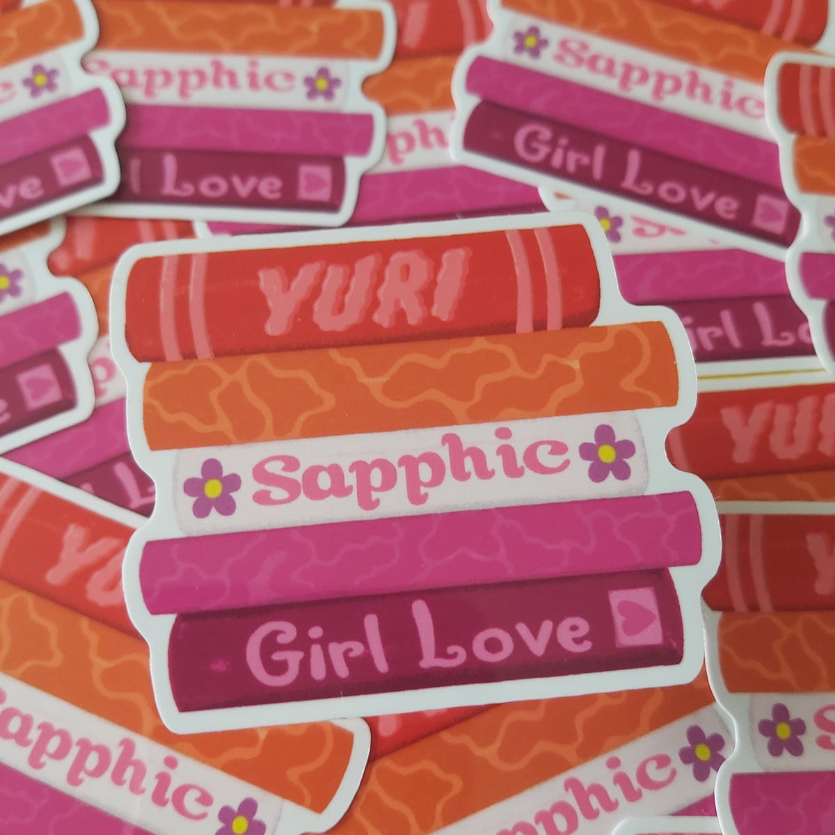 A pile of die-cut stickers featuring retro-style stacked book covers in pink, orange, and red, with text including 'YURI', 'Sapphic', and 'Girl Love'.