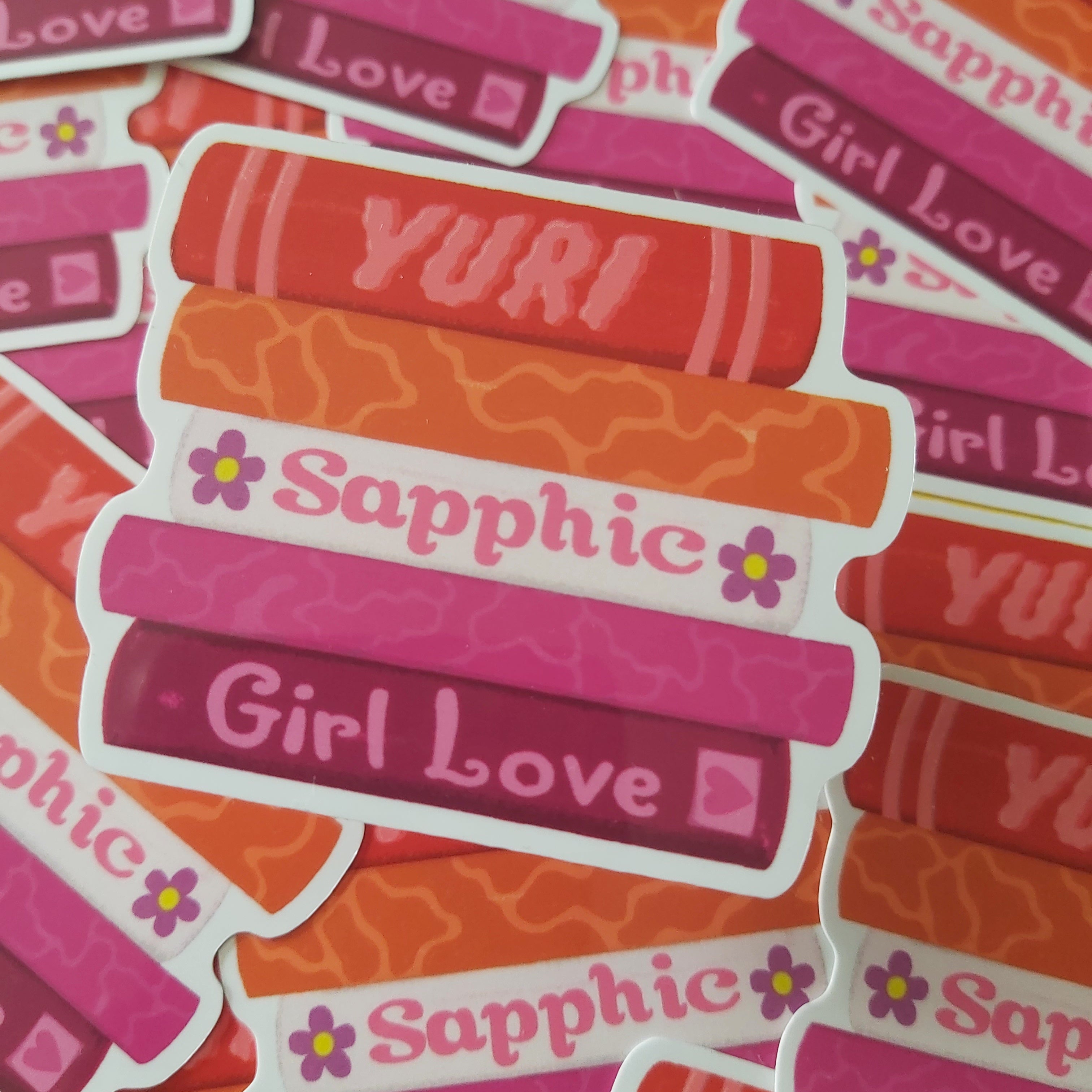 A pile of die-cut stickers featuring a striped book design in shades of pink, orange, and red, with the words 'Sapphic', 'Girl Love', and 'YURI' printed on them.