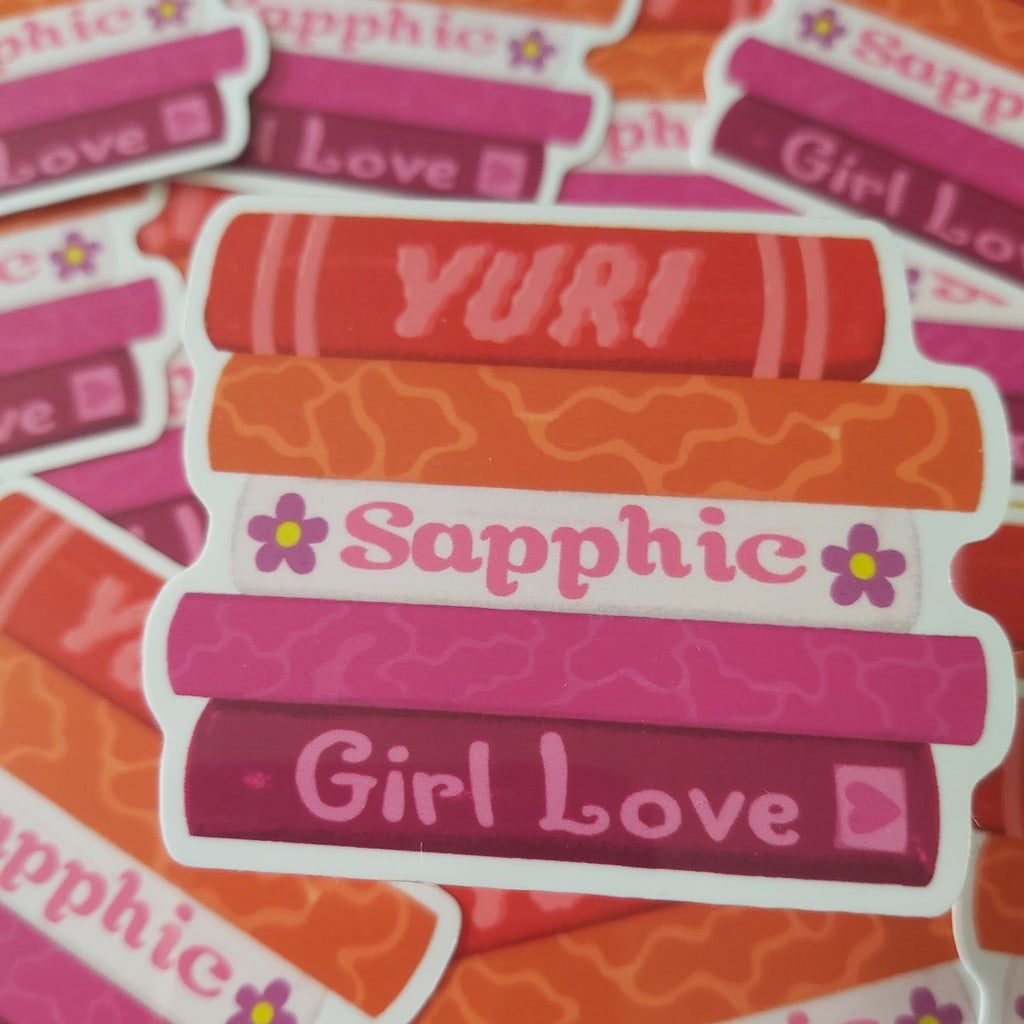A close-up pile of die-cut stickers featuring stacked book covers in shades of orange, pink, and red, with the words 'Sapphic', 'YURI', and 'Girl Love' written on them.