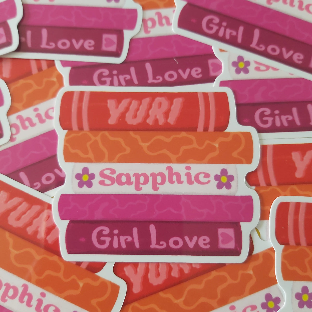 A pile of die-cut stickers featuring retro book covers in shades of orange, pink, and magenta, with white borders and text reading 'Sapphic', 'Girl Love', and 'YURI'.