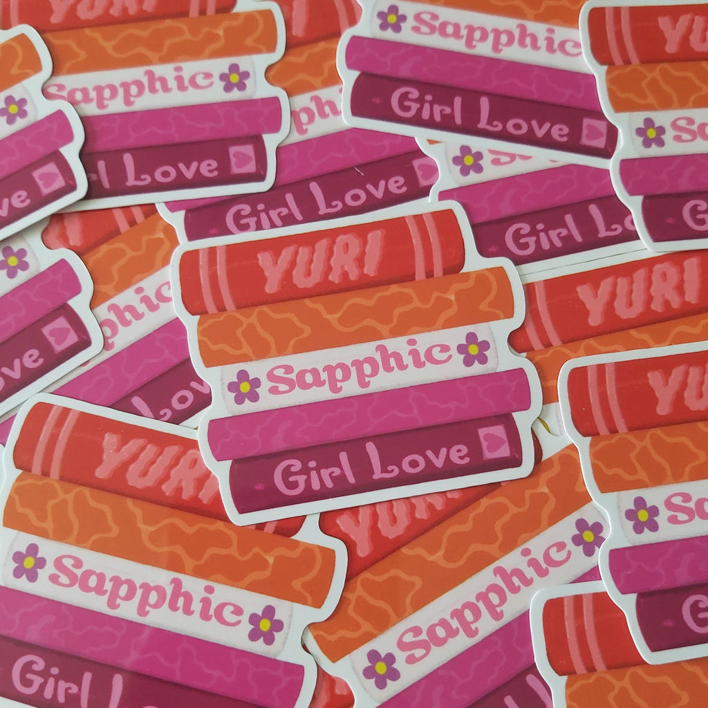 A pile of die-cut stickers featuring the phrase 'Girl Love' in white text on a pink background, with 'YURI' printed on orange-red patterned book covers above it. The stickers have white borders and small purple flower accents, photographed on a bright yellow background.
