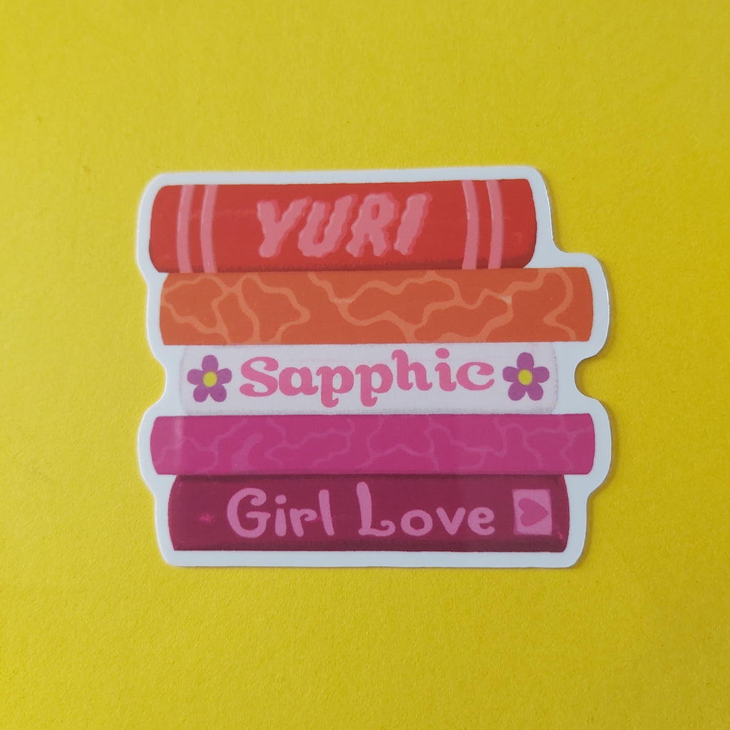 A die-cut sticker featuring five stacked books in shades of red, orange, and pink, with the words 'YURI', 'Sapphic', and 'Girl Love' printed on them, set against a solid yellow background.