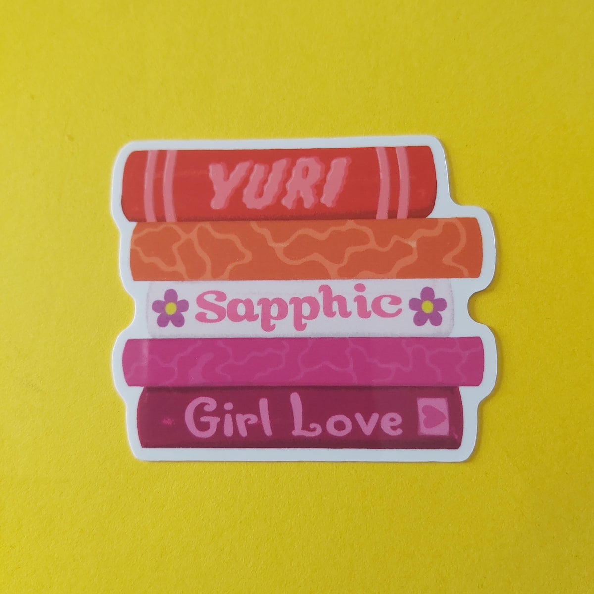 A die-cut sticker featuring five stacked books in shades of red, orange, and pink, with the words 'YURI', 'Sapphic', and 'Girl Love' printed on them, set against a solid yellow background.