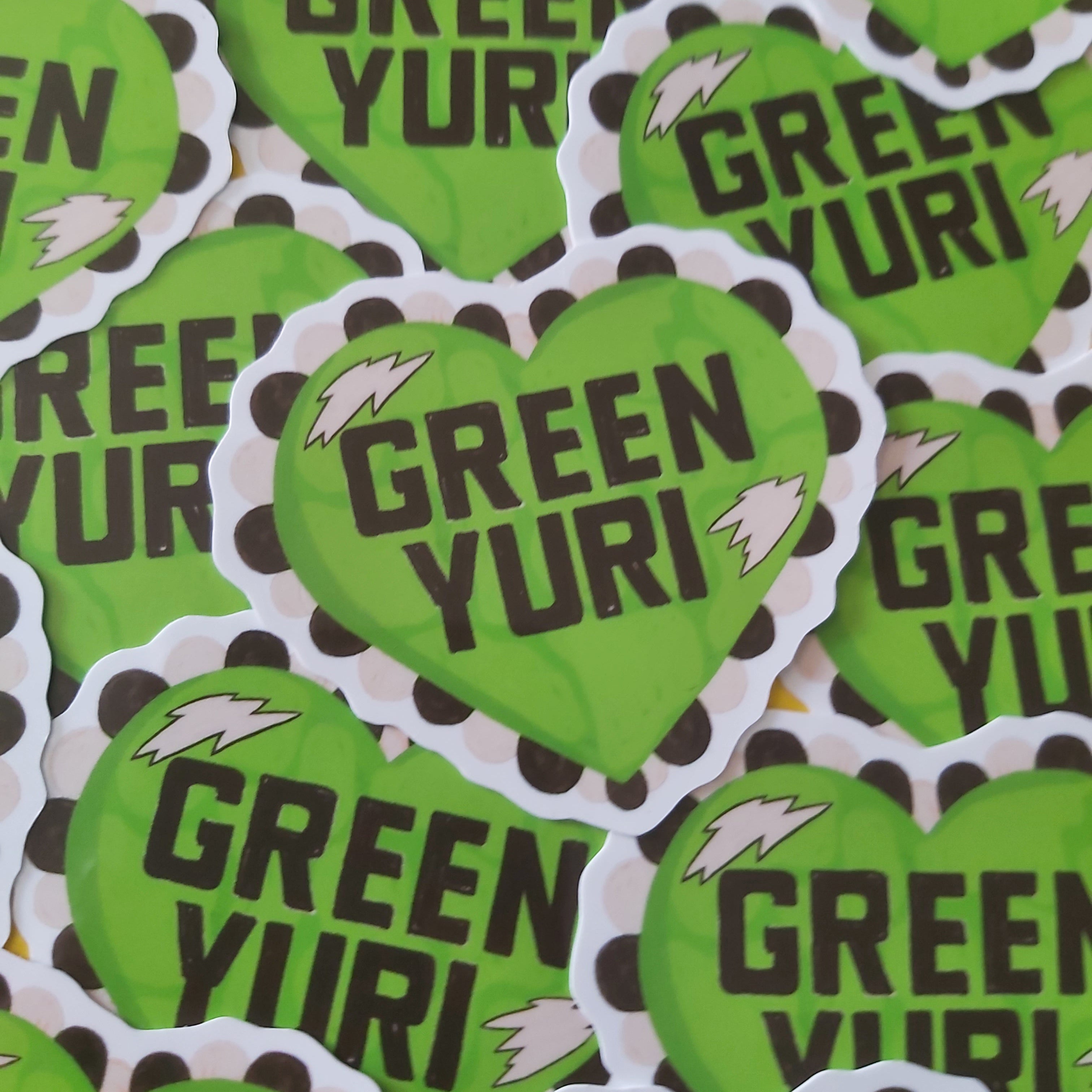 A pile of die-cut heart-shaped stickers featuring the text 'GREEN YURI' in bold black letters on a bright green background, with a scalloped border around the edges and small white lightning bolt accents.