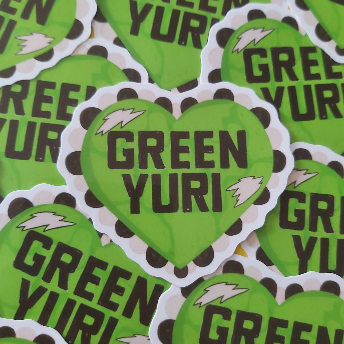A pile of die-cut heart-shaped stickers featuring the text 'GREEN YURI' in dark brown lettering on a bright lime green background, with a white border and small white accents.