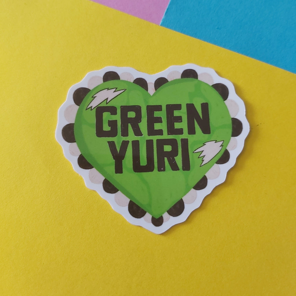 A heart-shaped sticker with a green centre, black and white scalloped border, and the words 'GREEN YURI' in black text, placed on a yellow background with pink and blue sections in the upper corners.