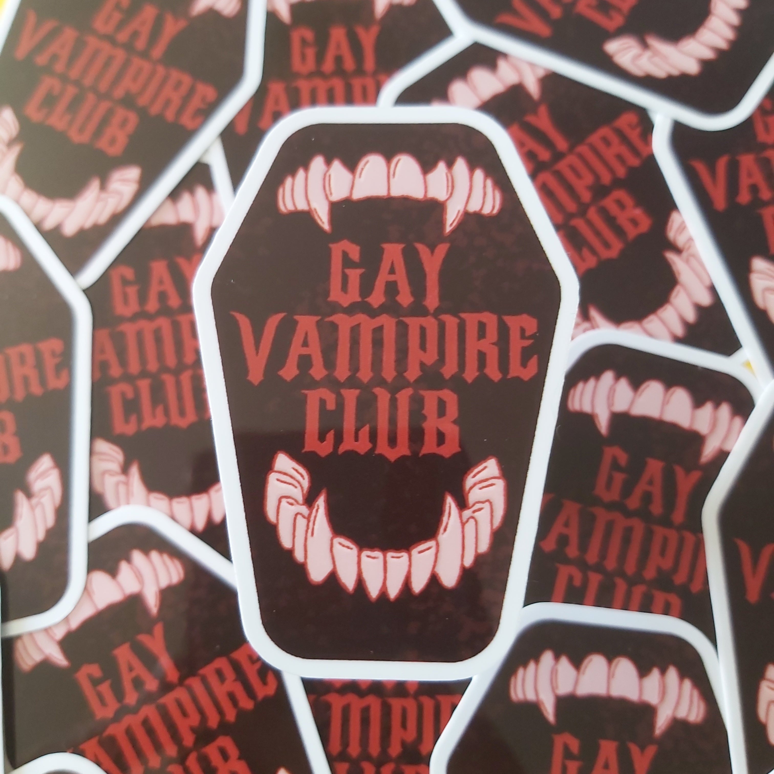 A pile of die-cut stickers featuring a coffin-shaped design with the words 'GAY VAMPIRE CLUB' in red over a dark brown background, with off-white vampire fangs at the top and bottom.