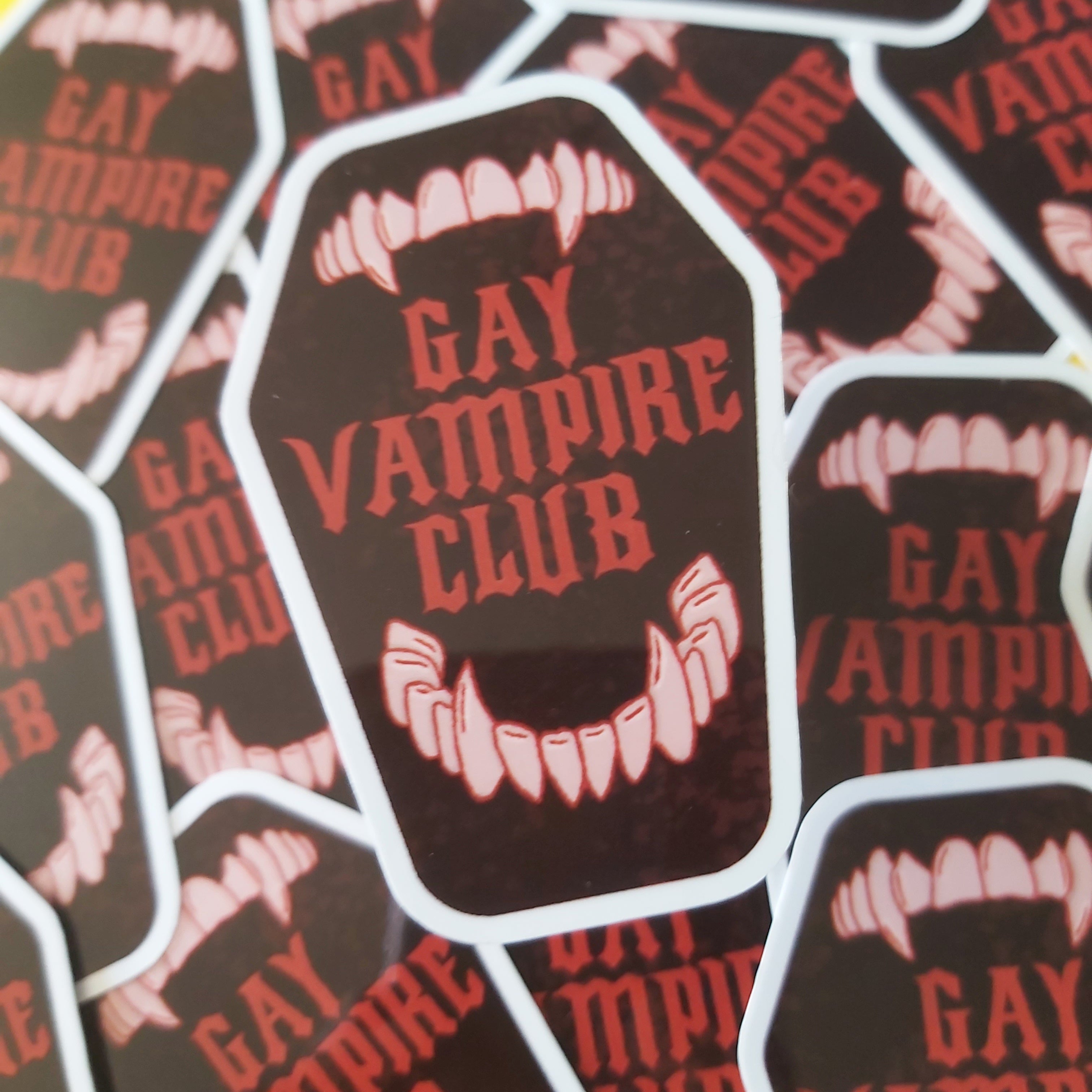A close-up of multiple identical die-cut stickers. Each sticker is dark brown with red text reading 'GAY VAMPIRE CLUB' and white vampire fangs and border details.