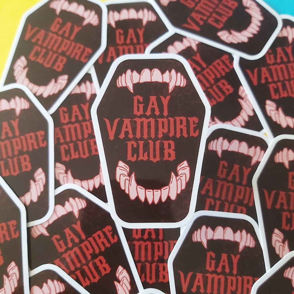 A pile of die-cut stickers shaped like coffins, featuring the text 'GAY VAMPIRE CLUB' in red lettering over a dark brown background, with white vampire fangs illustrated above and below. The stickers are scattered on a brightly colored background with yellow, pink, and blue sections.
