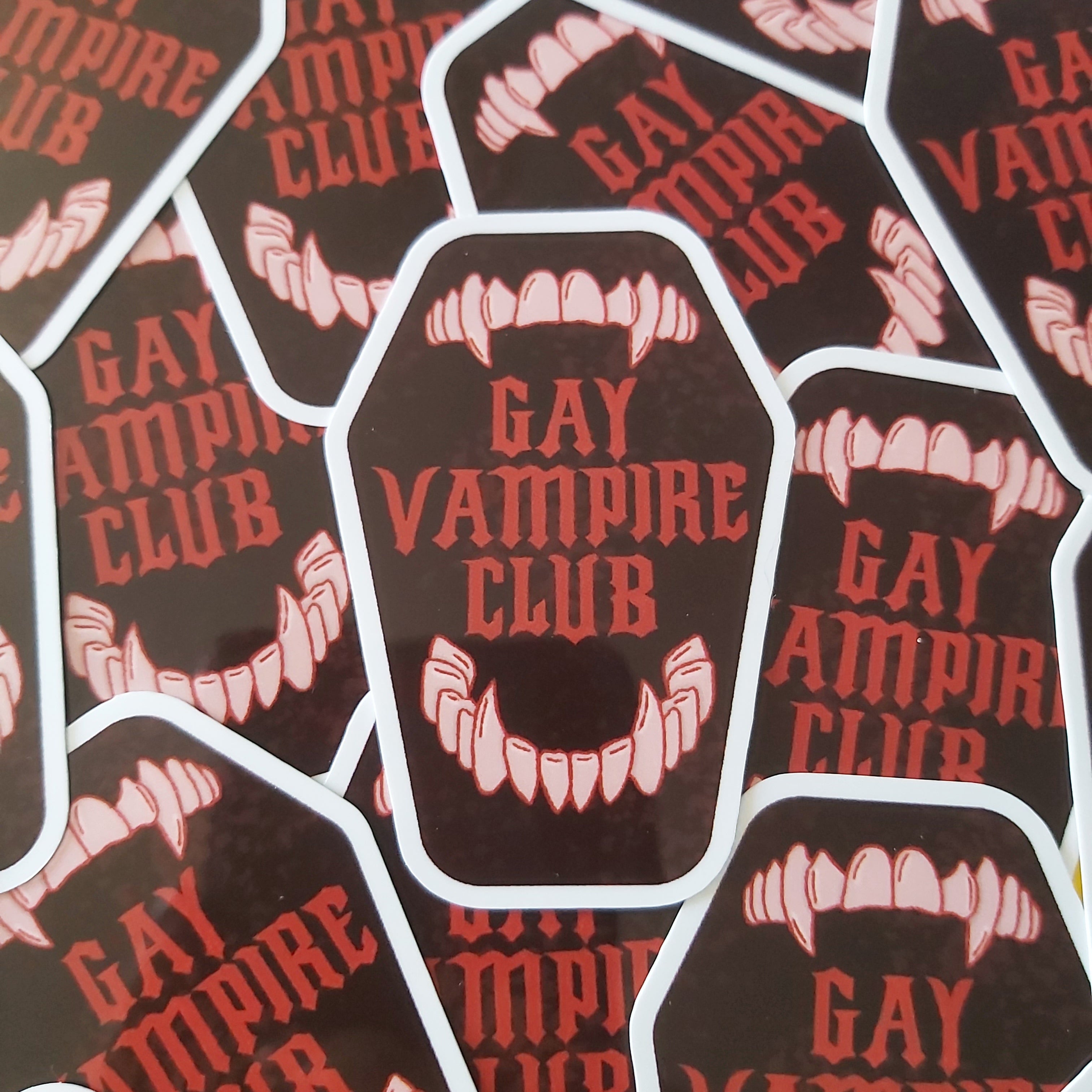 A pile of die-cut stickers featuring a dark brown coffin-shaped design with the words 'GAY VAMPIRE CLUB' in red and a vampire mouth graphic with white teeth.