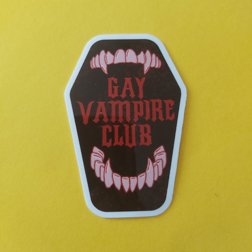 A coffin-shaped sticker with the words 'GAY VAMPIRE CLUB' in red gothic lettering, featuring off-white vampire fangs at the top and bottom, on a bright yellow background.