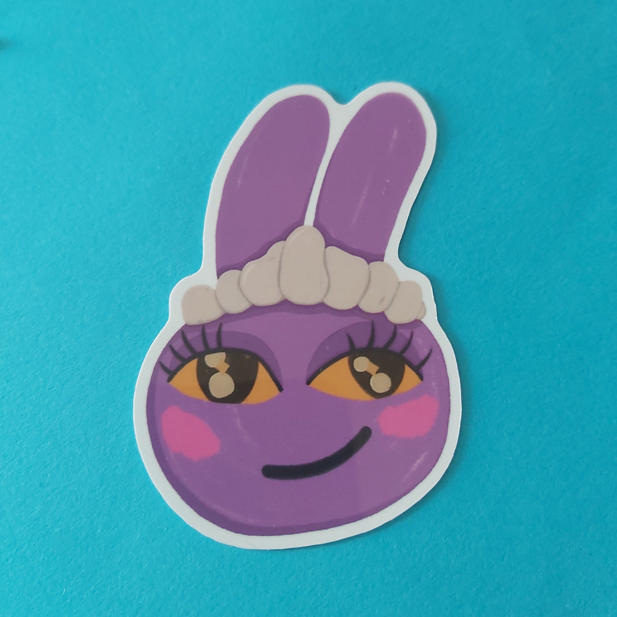 A die-cut sticker featuring a purple cartoon bunny face with long ears called Jax, a white maid headband, and yellow eyes, shown on a solid bright blue background.