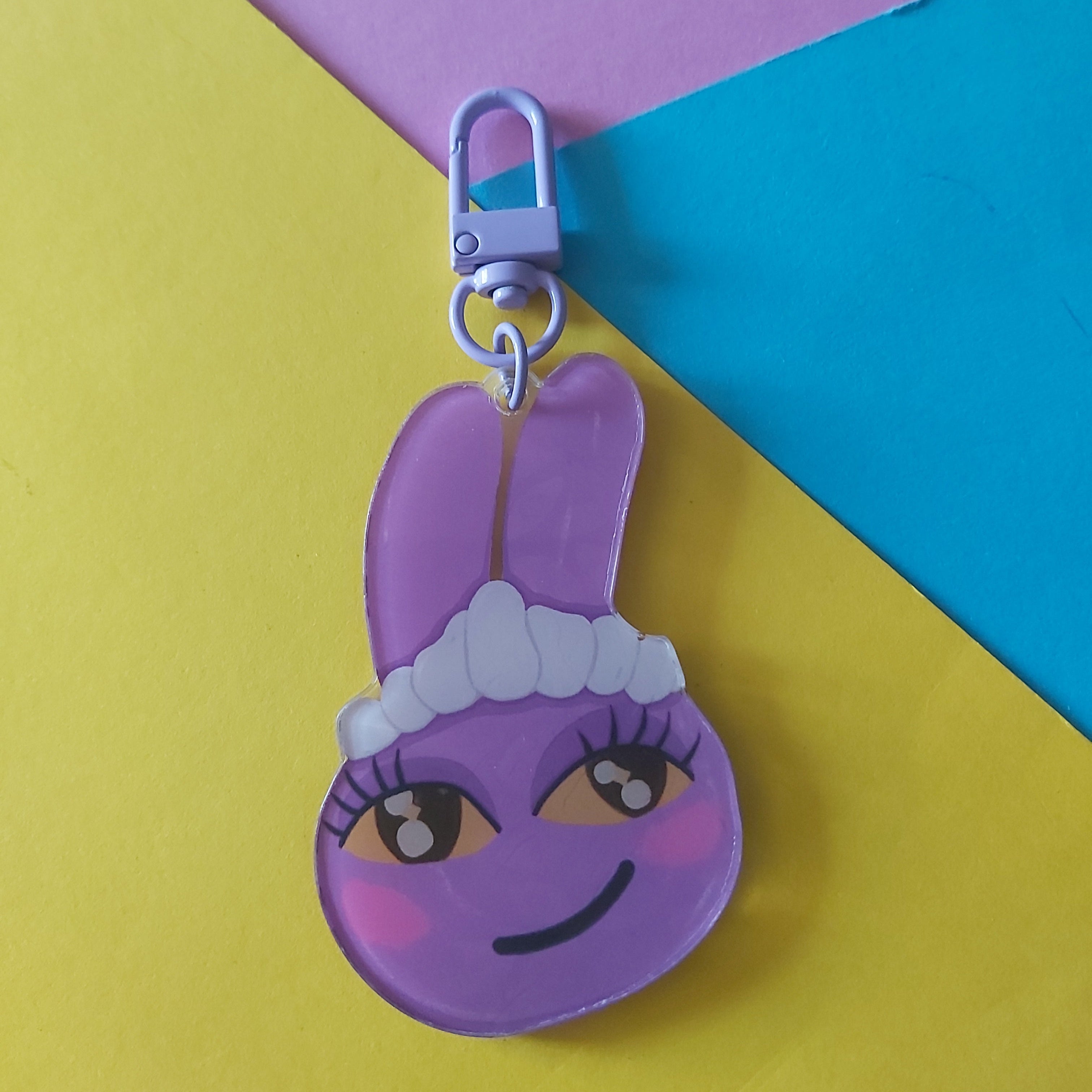 A purple cartoon bunny keychain charm with a silver lobster clasp, resting on a diagonally split yellow, blue, and purple background.