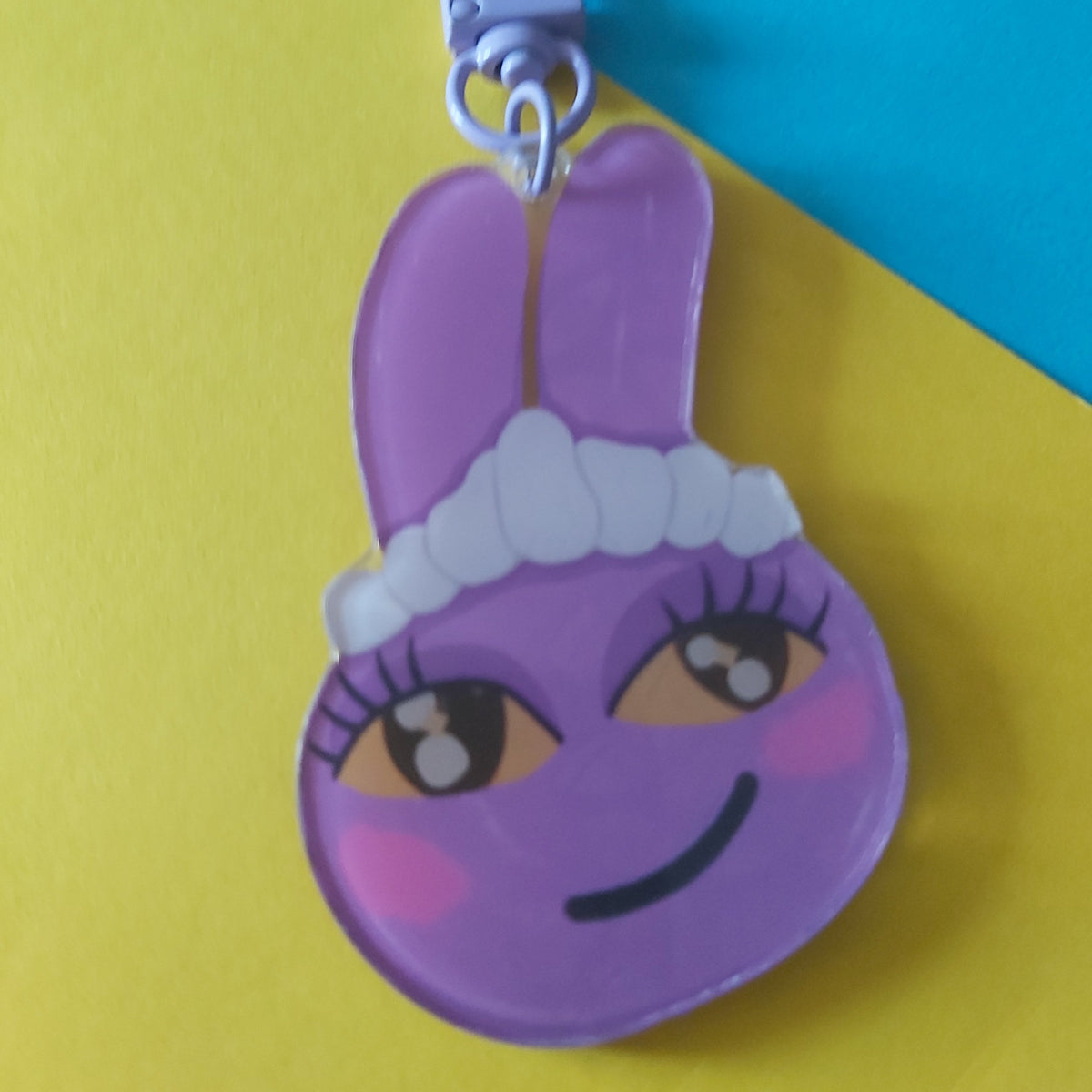 A purple bunny-shaped keychain charm with a cartoon face, pink cheeks, and a white headband, attached to a lilac metal clasp, set against a colourful background of yellow, blue, and pink paper.