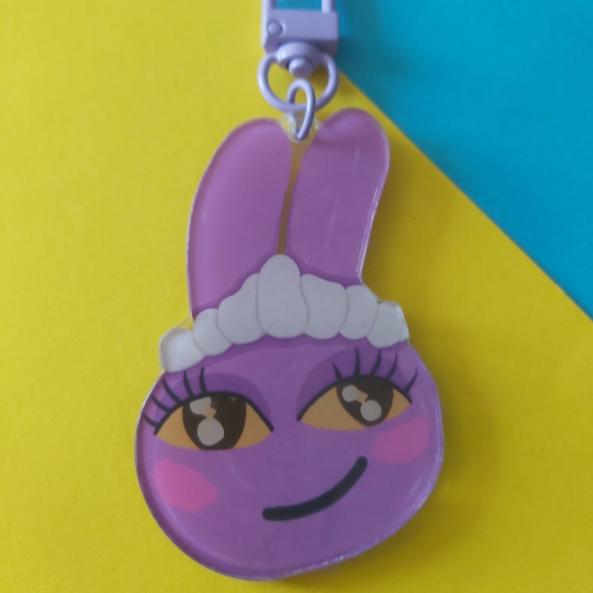 A purple cartoon bunny keychain charm with a purple lobster clasp, resting on a colourful background of yellow, pink, and blue paper.