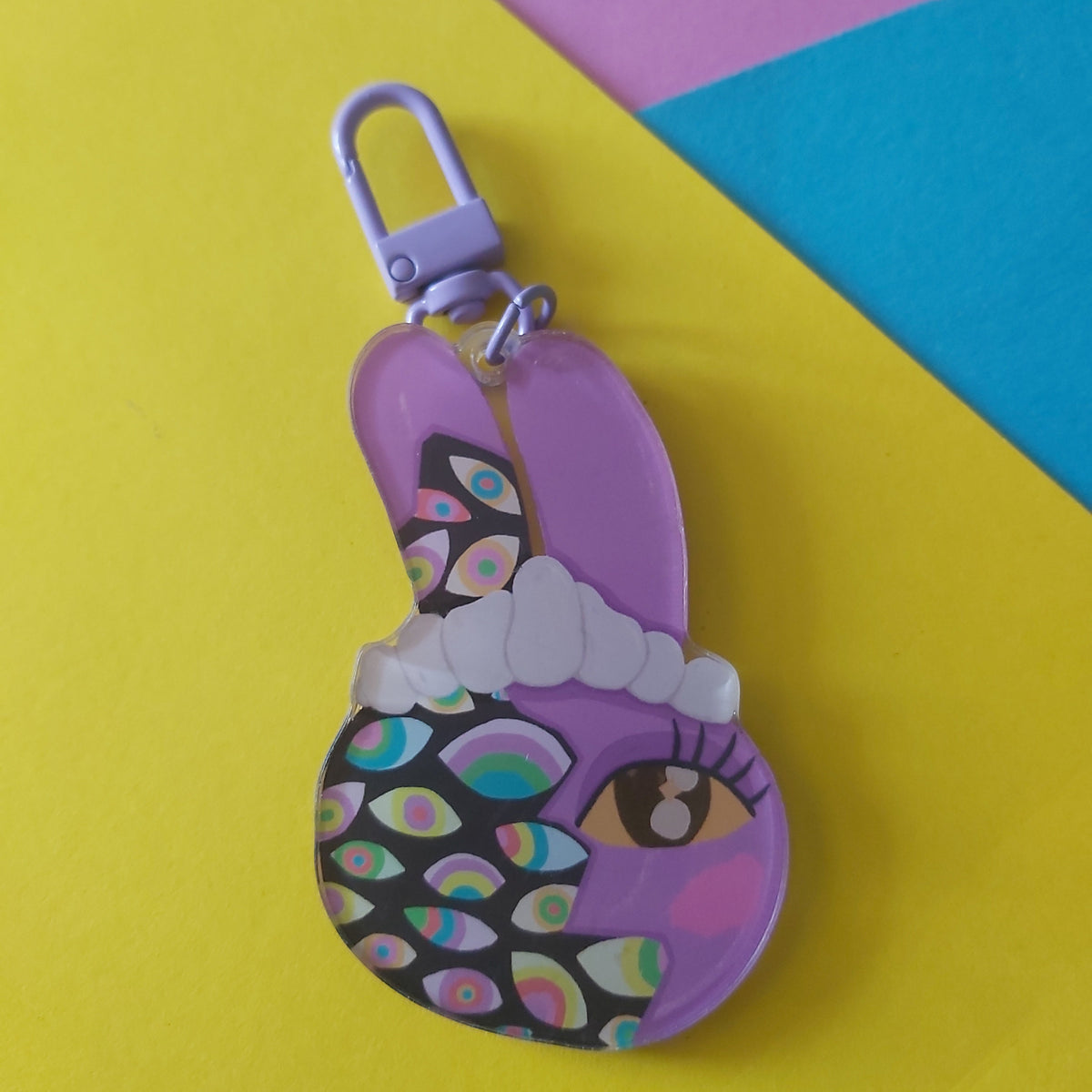 A purple bunny-shaped keychain or bag charm with a lobster clasp, featuring a stylised face with a black patterned section of colourful eye details and a white headband, displayed on a diagonally split yellow, pink, and blue background.