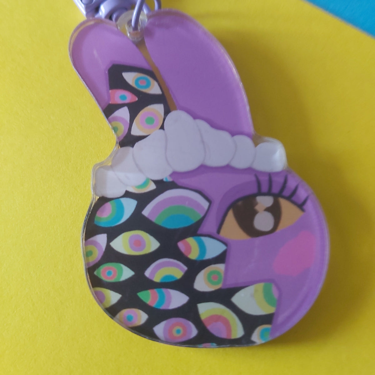 A purple bunny-shaped keychain or charm with a black face patterned with colourful rainbow eyes, a white headband, and a purple clasp at the top, shown on a bright yellow background with a blue corner.