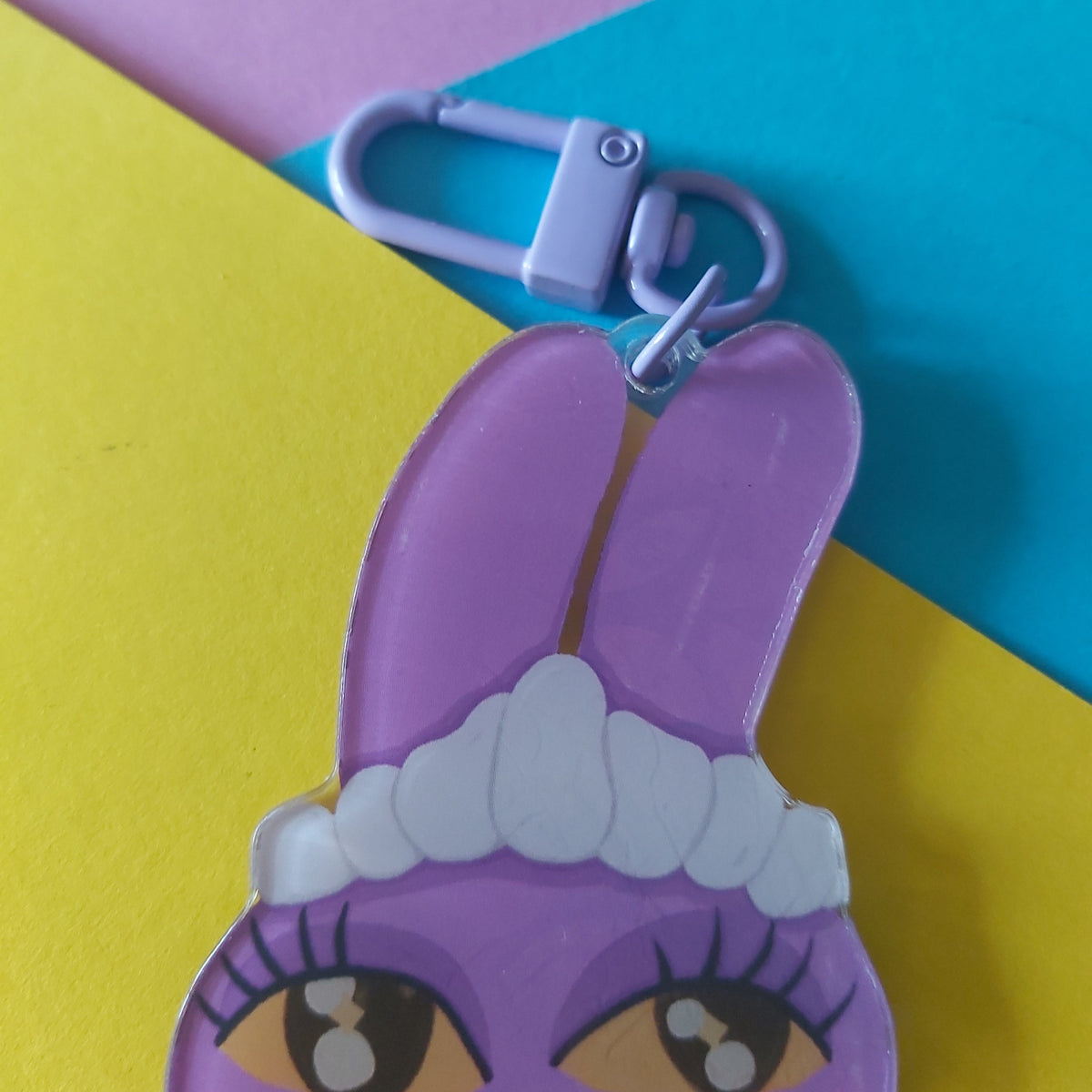 A close-up of a purple cartoon bunny keychain charm with long ears, yellow eyes, and a white maid headband, attached to a lavender metal clasp. The charm is photographed on a colourful background with pink, blue, and yellow paper sections.