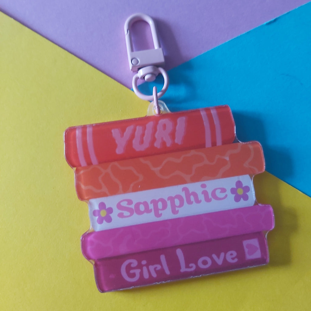 A colorful acrylic keychain charm shaped like a stack of books with the names 'YURI', 'Sapphic', and 'Girl Love' written on them, attached to a silver lobster clasp. The charm is displayed on a diagonally split yellow and purple background with a blue section.