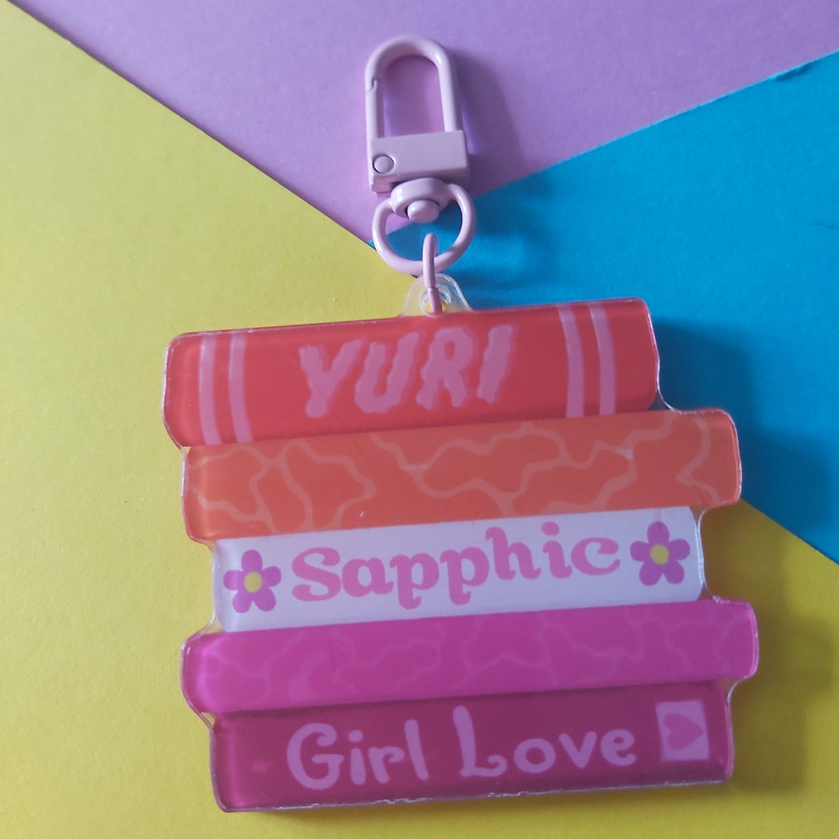 A colourful acrylic keychain charm shaped like a stack of books with the words 'YURI', 'Sapphic', and 'Girl Love' printed on them, attached to a silver lobster clasp. The background is a vibrant, diagonally split yellow, purple, and blue surface.