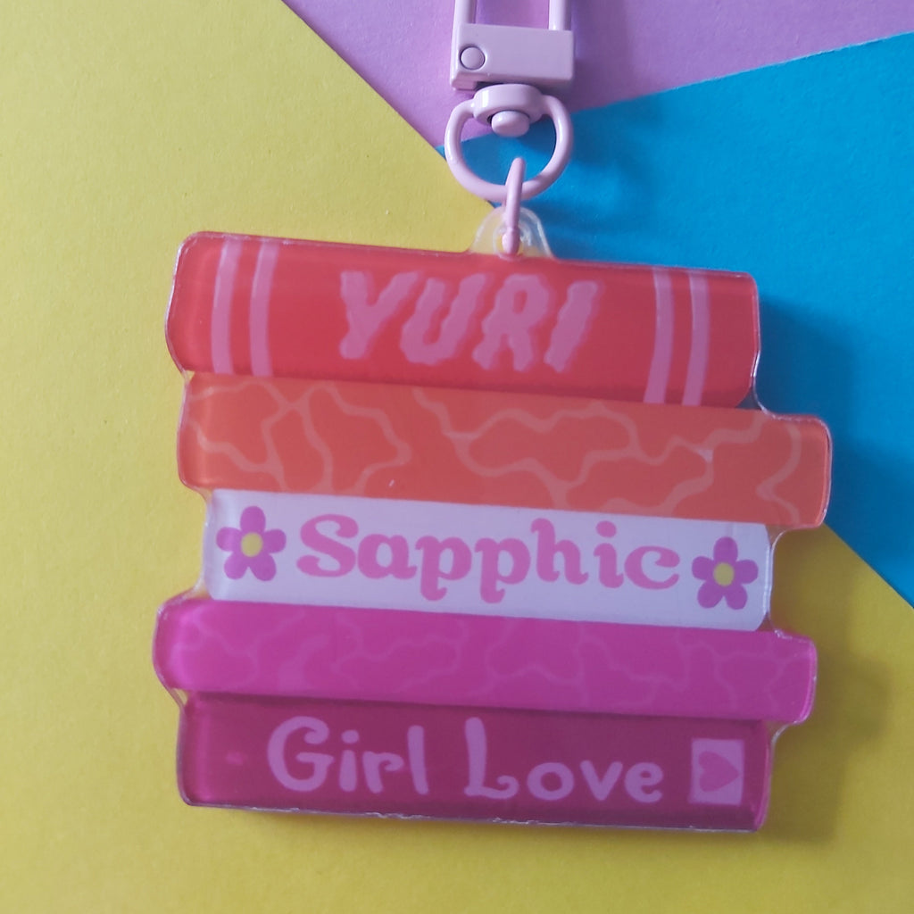 A colorful keychain charm shaped like a stack of five books with the words 'YURI', 'Sapphic', and 'Girl Love' printed on them, attached to a pink metal lobster clasp. The background is a geometric colour-blocked paper design in purple, yellow, and blue.