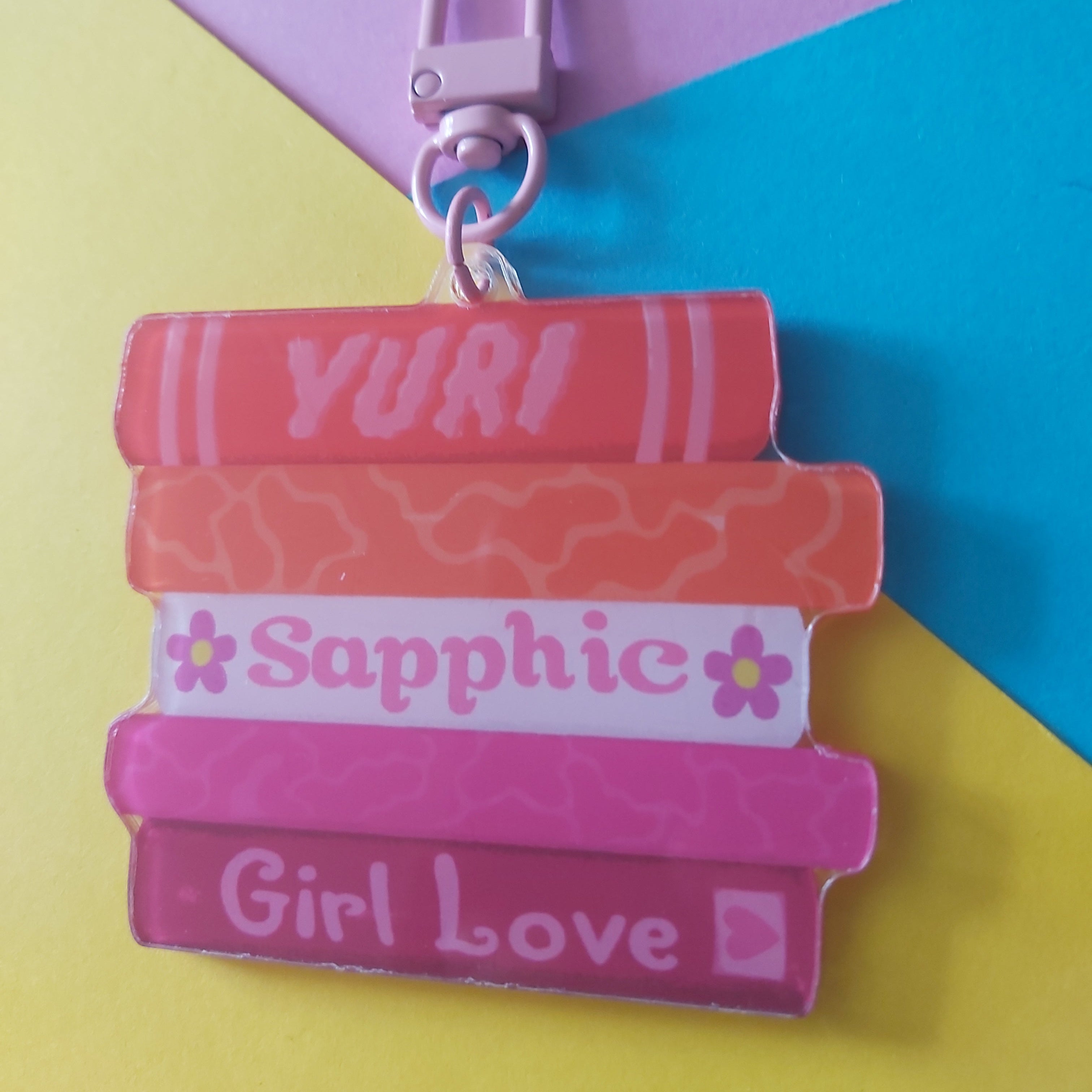 A stack of book-shaped charms with the words 'YURI', 'Sapphic', and 'Girl Love' printed on them, attached to a light pink clasp, set against a colourful background of yellow, blue, and pink paper.