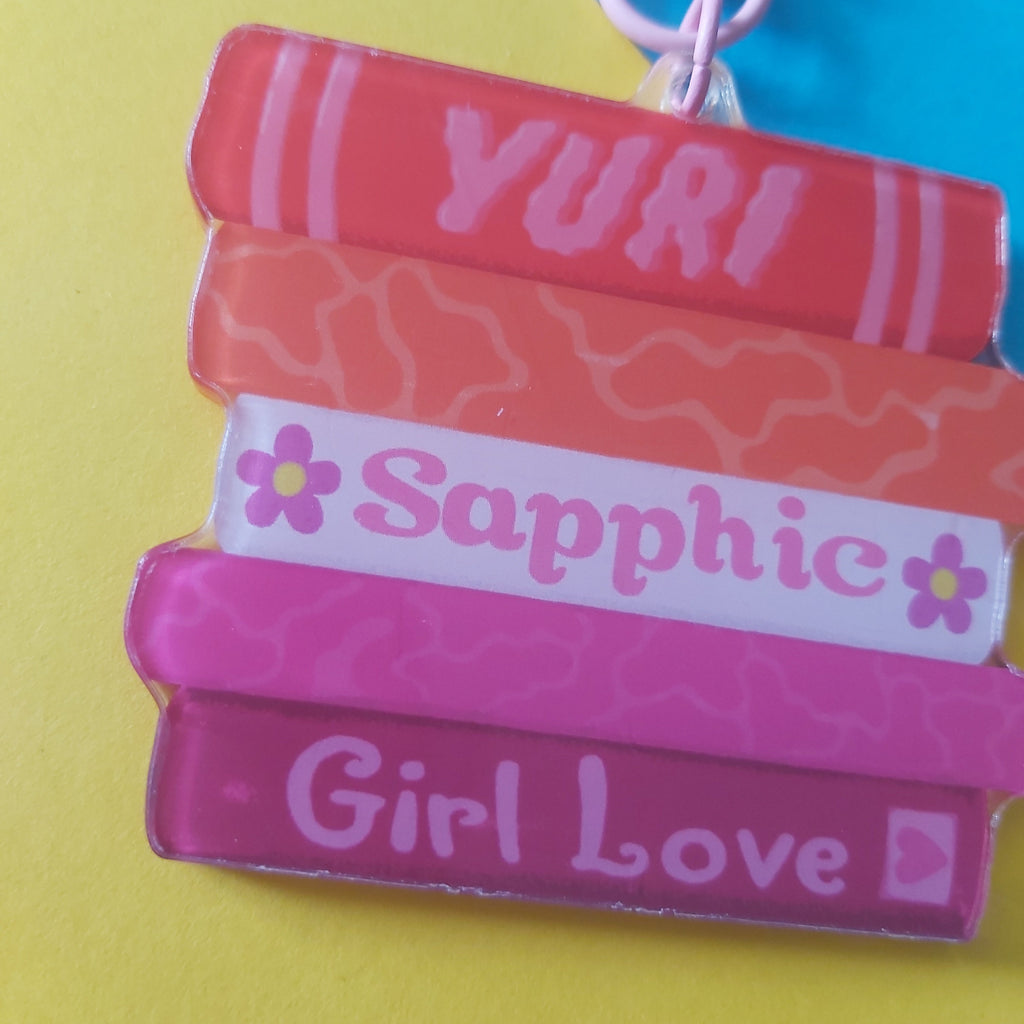 A close-up of a colorful acrylic keychain shaped like a stack of books with the words 'YURI', 'Sapphic', and 'Girl Love' written on them, attached to a pink clasp against a bright yellow background.