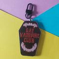 A coffin-shaped keychain bag charm with a black carabiner clip, featuring the text 'GAY VAMPIRE CLUB' in red between white vampire fang illustrations, set against a diagonally split yellow, turquoise, and purple background.