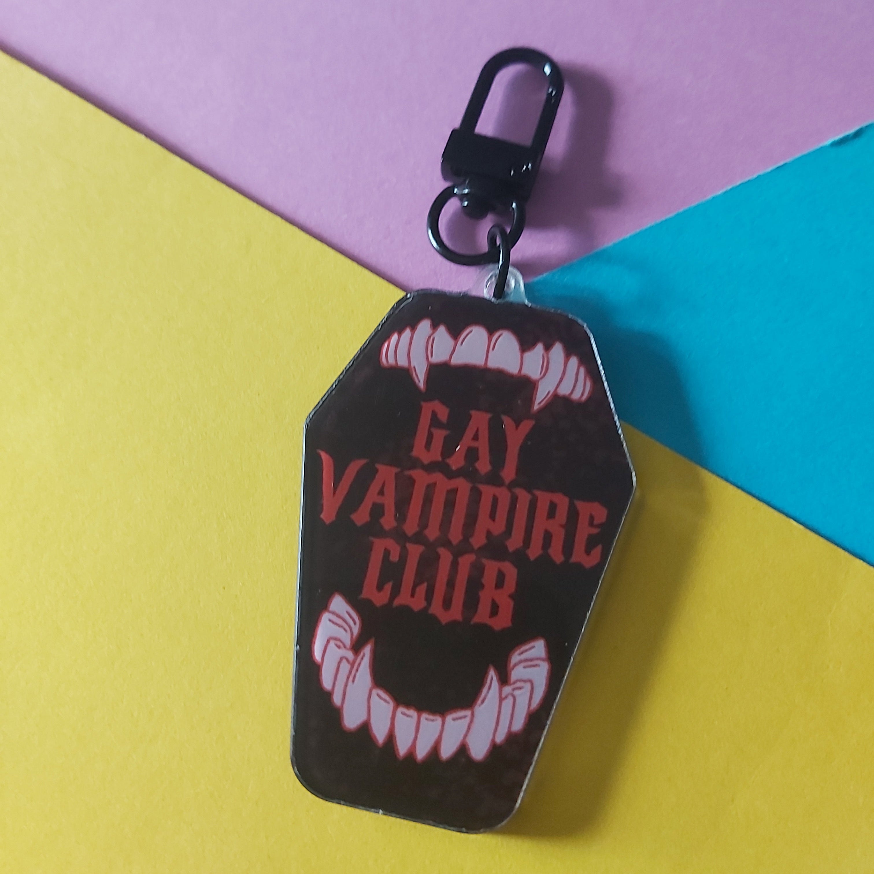 A coffin-shaped keychain bag charm with a black clasp, featuring a dark brown background with red text reading 'GAY VAMPIRE CLUB' and white vampire fangs at the top and bottom, displayed on a yellow, pink, and blue geometric background.