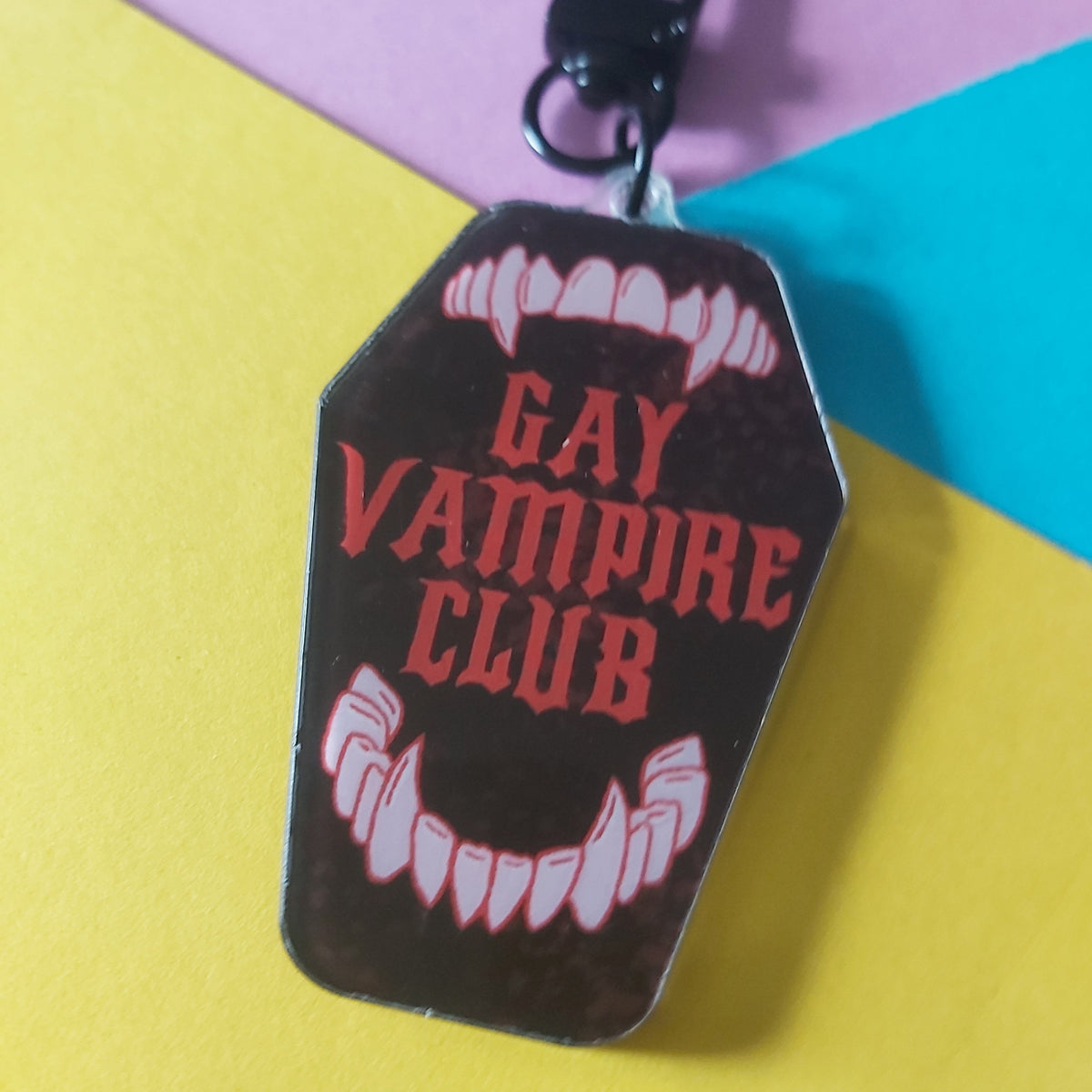 A coffin-shaped keychain or bag charm with a black clasp, featuring the words 'GAY VAMPIRE CLUB' in red text between white vampire fangs, displayed on a colourful yellow, pink, and blue background.