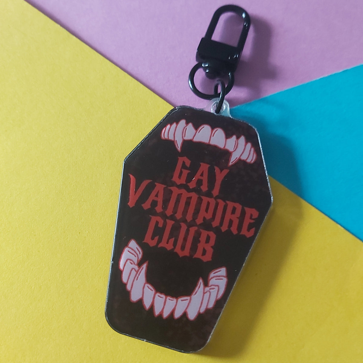 A coffin-shaped keychain or bag charm with a black clasp, featuring the words 'GAY VAMPIRE CLUB' in red text over white vampire teeth graphics on a dark brown background, set against a colourful yellow, purple, and blue geometric backdrop.