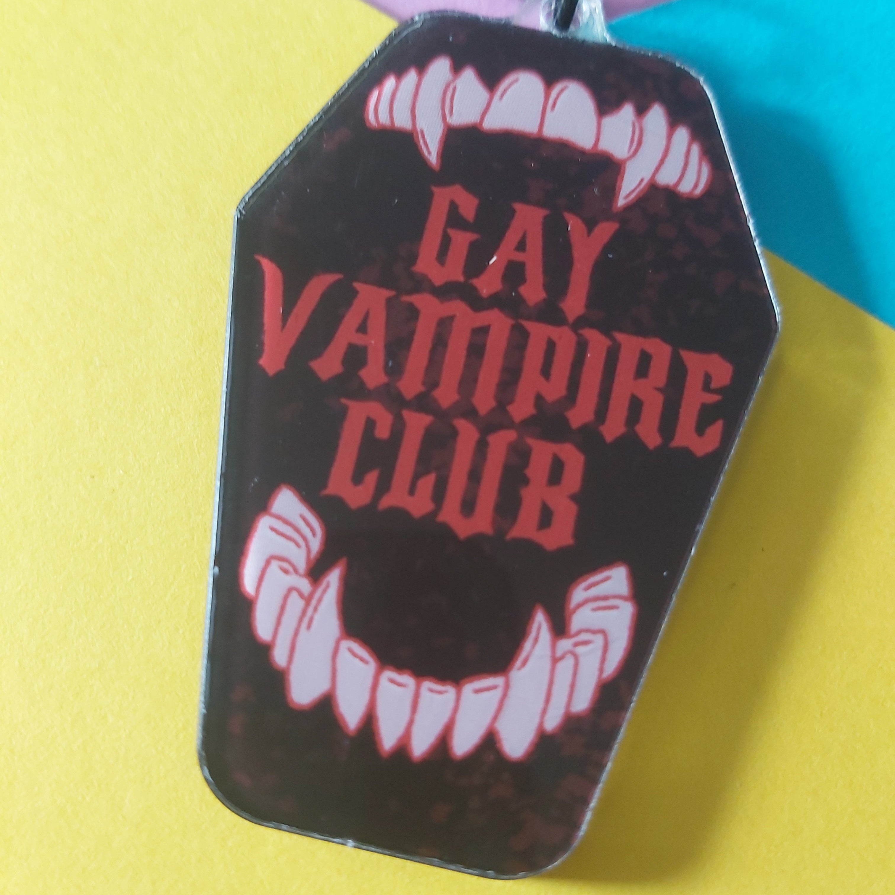 A coffin-shaped keychain with a black background, red gothic text reading 'GAY VAMPIRE CLUB,' and white vampire fangs and mouth details, photographed on a bright yellow surface with colourful paper accents in the background.