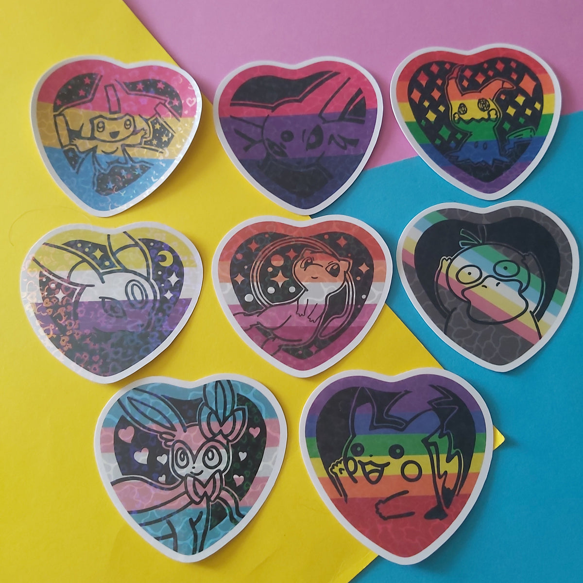 Seven heart-shaped die-cut stickers featuring colourful cartoon Pokémon illustrations in pride flag backgrounds, arranged on a diagonally split yellow and pink background.