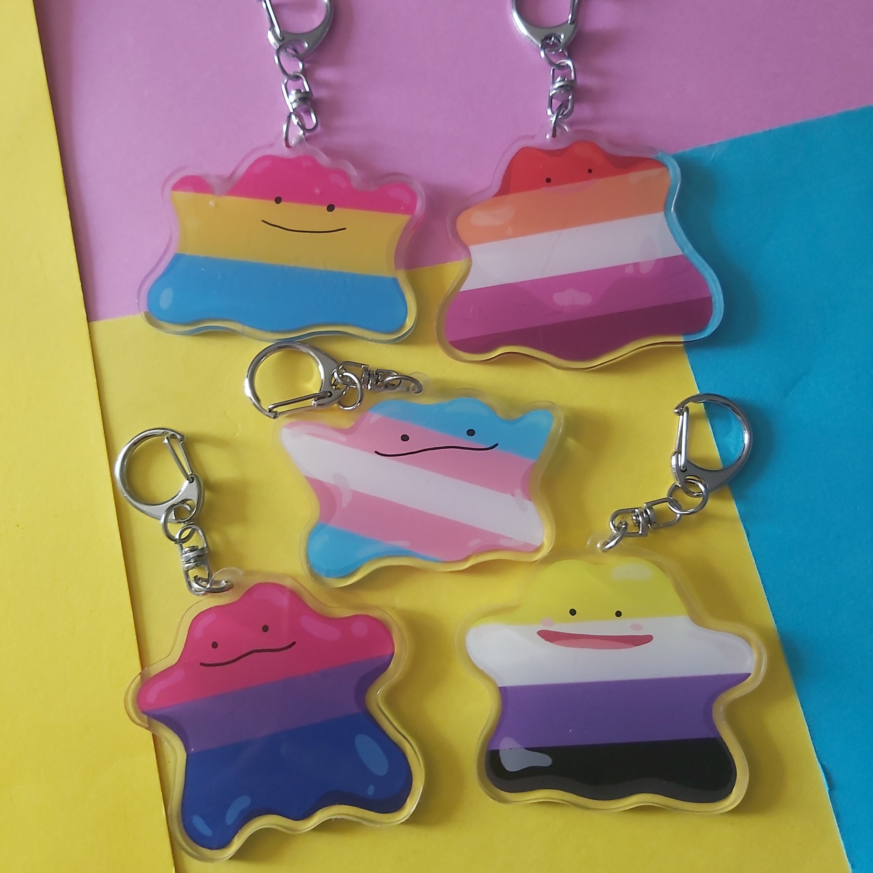 Five pride-flag-striped ditto keychains with silver lobster clasps, arranged on a bright pink, yellow, and blue background.