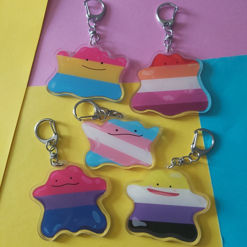 Five pride flag keychain charms shaped ditto characters, arranged on a colourful yellow, pink, and blue background.