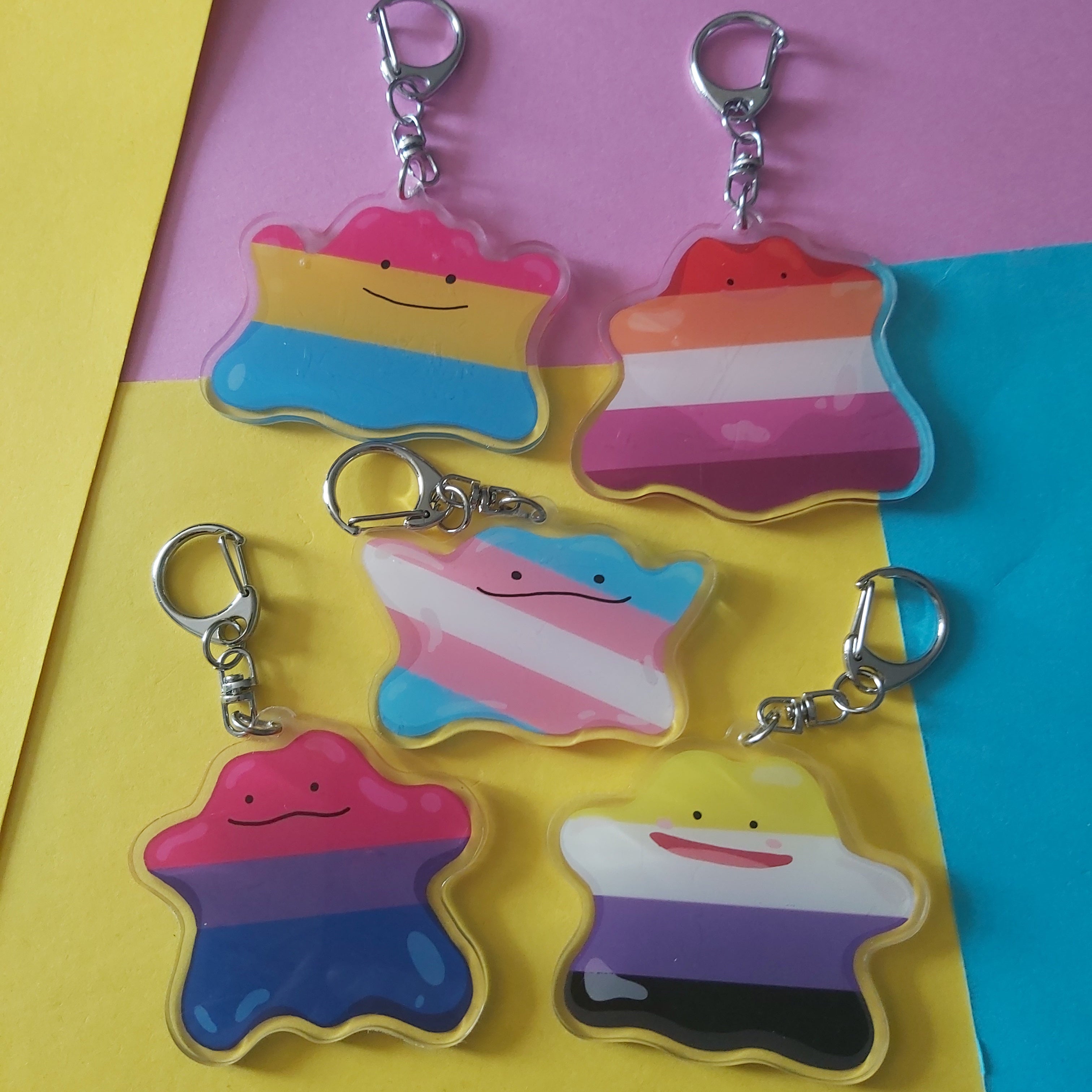 Five pride flag keychain charms shaped ditto characters, arranged on a colourful yellow, pink, and blue background.