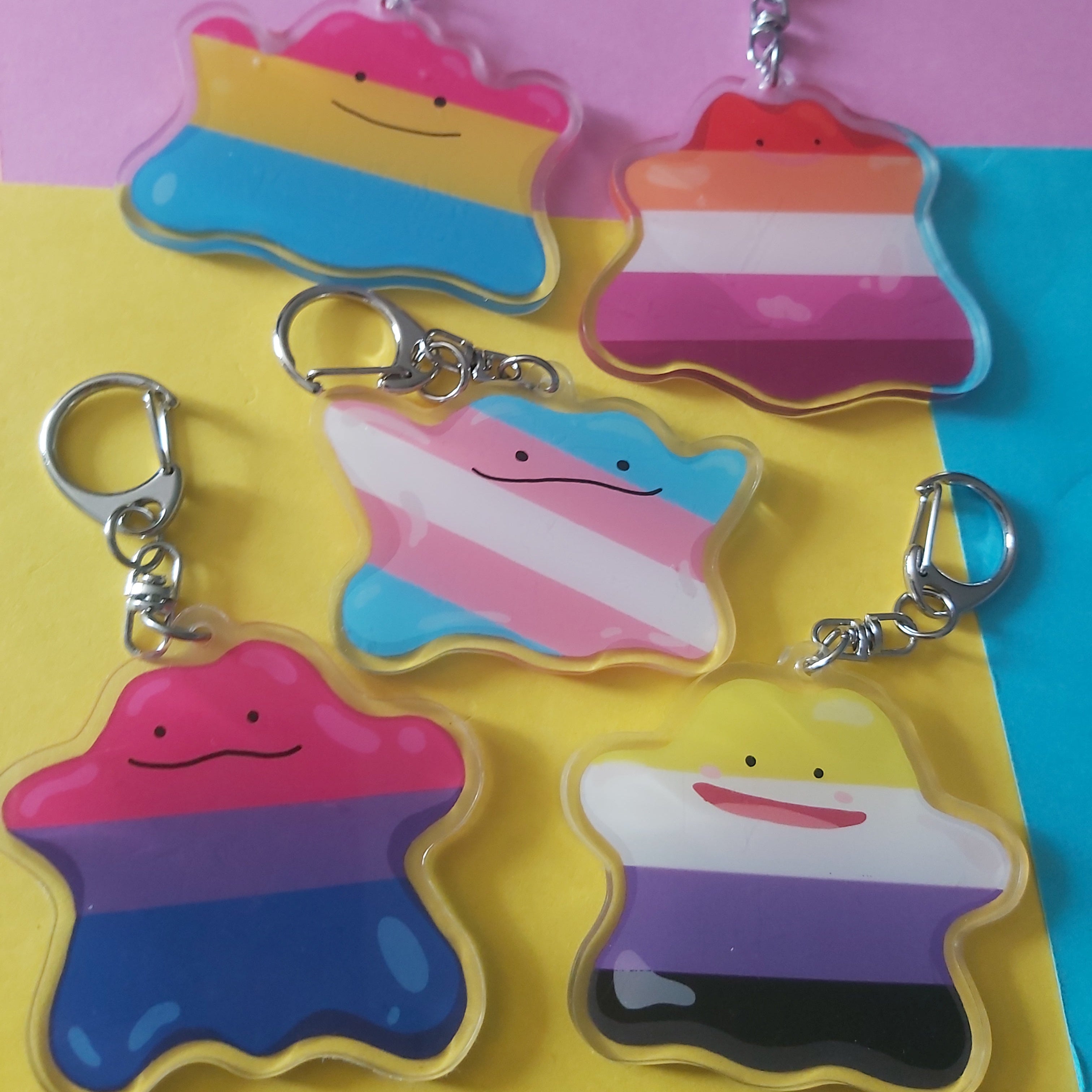 Five acrylic keychains with silver keyrings, each featuring a different striped pride flag design, arranged on a colourful pastel background.