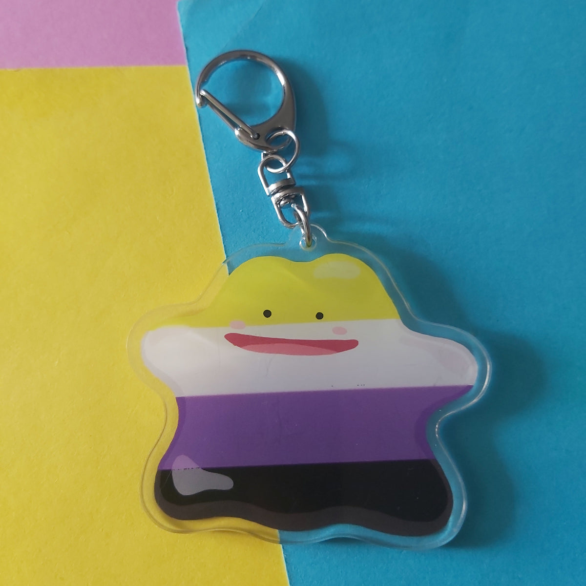 A non-binary pride keychain shaped like a blob character, featuring the stripes of purple, white, yellow, and black attached to a silver metal clasp. The keychain is displayed on a background split diagonally between yellow and blue paper with a pink corner visible.
